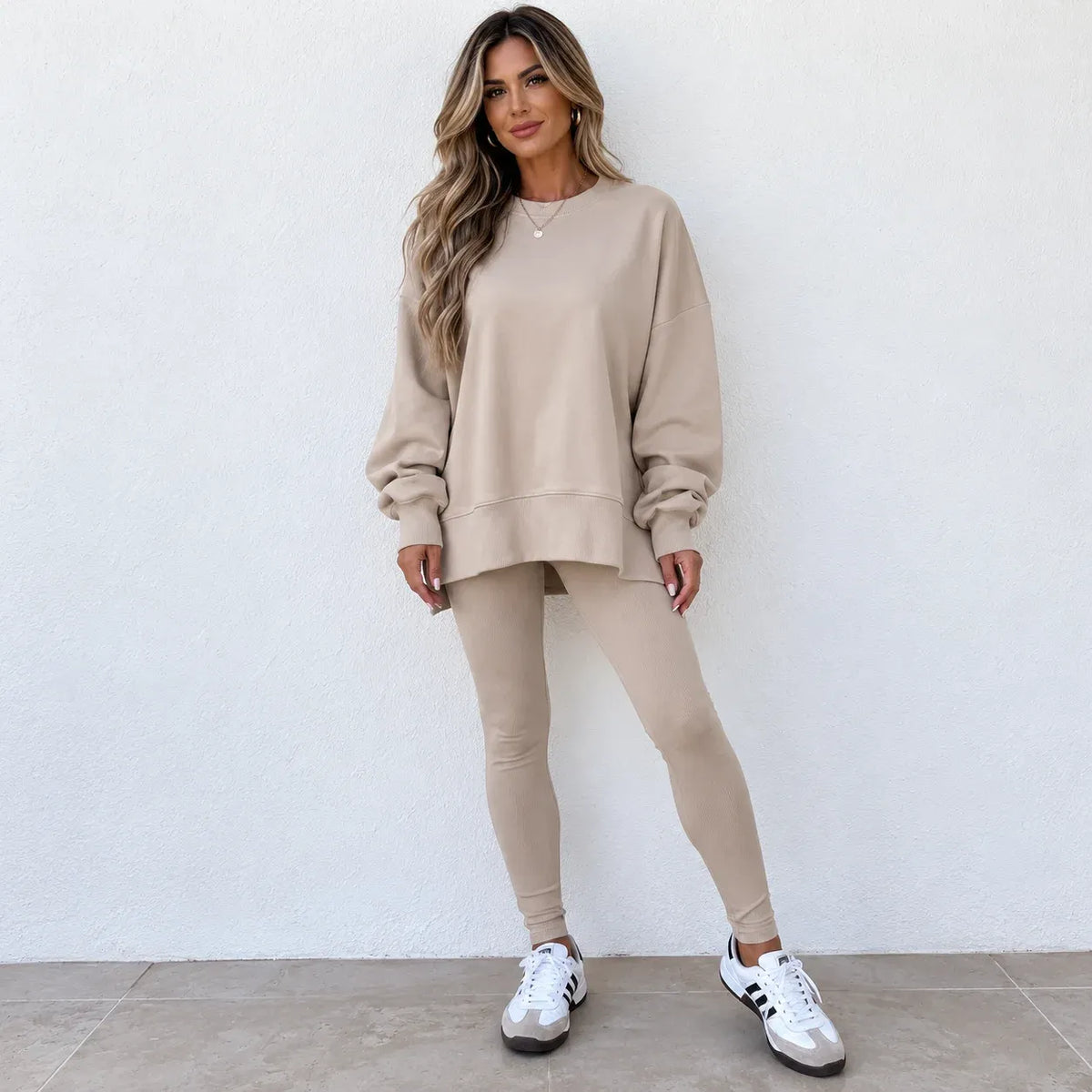 A woman modeling a matching beige lounge set consisting of a long-sleeve sweatshirt and high-waisted leggings, paired with white sneakers, standing against a white textured wall.