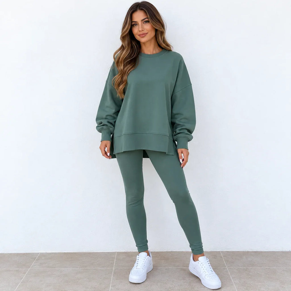 A woman modeling a matching sage green sweatshirt and leggings set, paired with white sneakers, standing against a plain white wall.