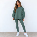 A woman modeling a matching sage green sweatshirt and leggings set, paired with white sneakers, standing against a plain white wall.