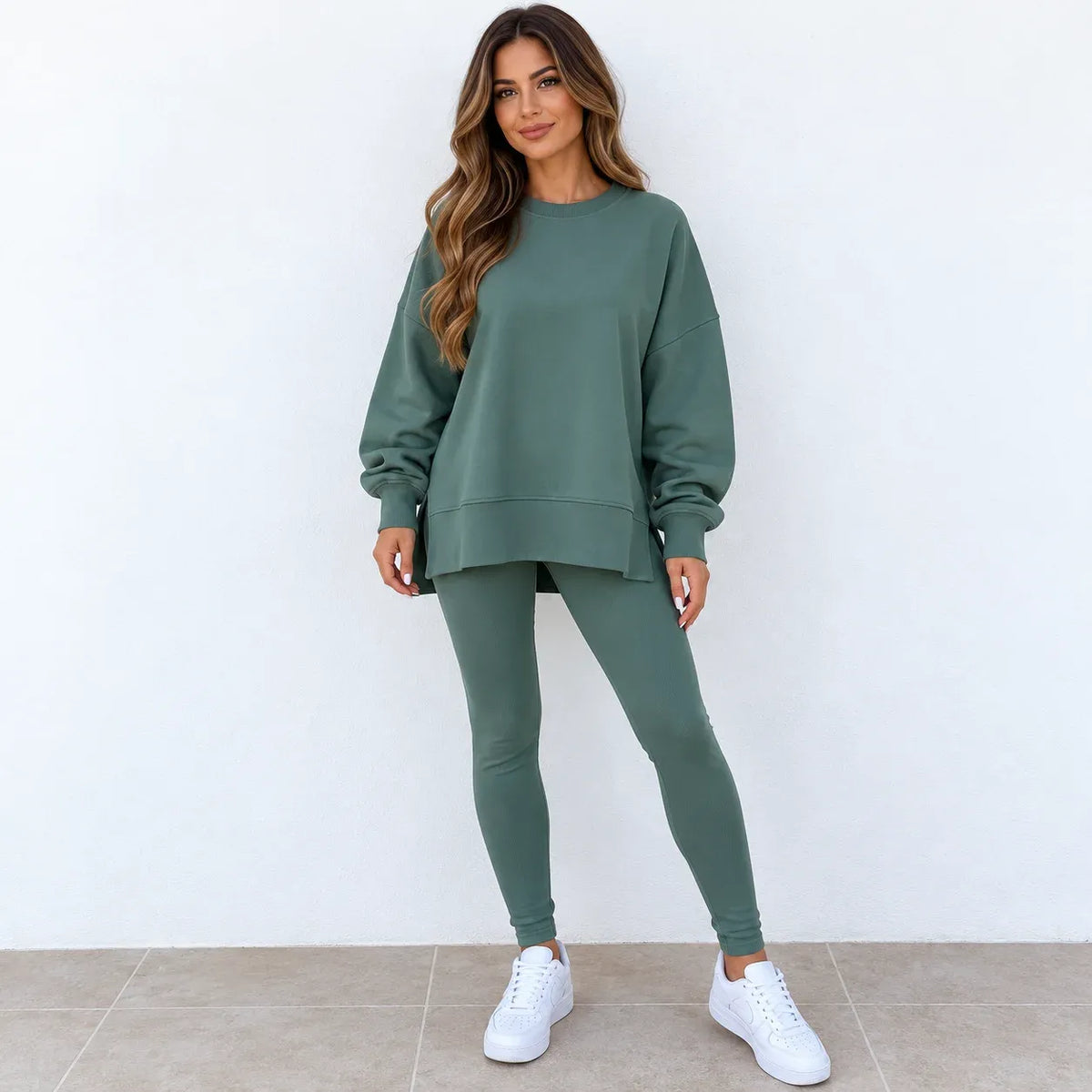A woman modeling a matching sage green sweatshirt and leggings set, paired with white sneakers, standing against a plain white wall.