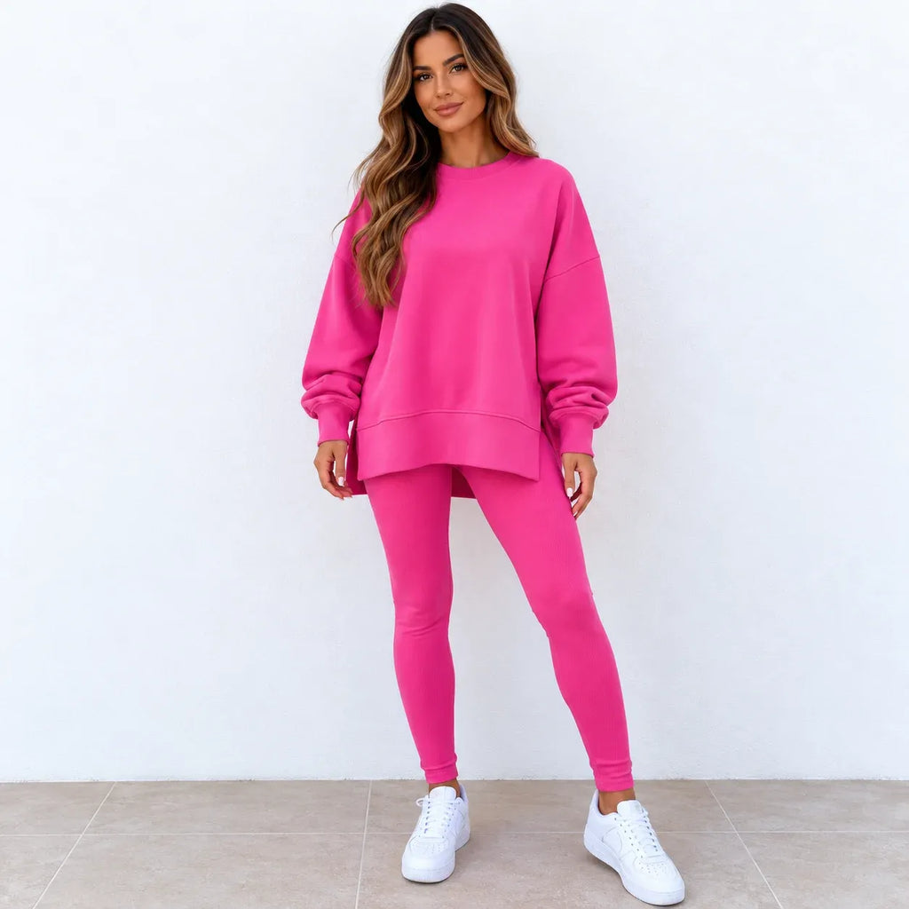 A woman modeling a bright pink two-piece lounge set consisting of an oversized crewneck sweatshirt and matching leggings, paired with white sneakers against a white wall and tiled floor.