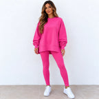 A woman modeling a bright pink two-piece lounge set consisting of an oversized crewneck sweatshirt and matching leggings, paired with white sneakers against a white wall and tiled floor.