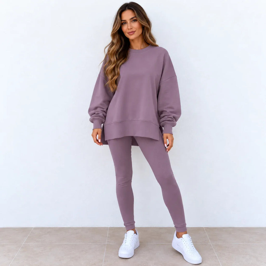 A woman modeling a matching dusty purple sweatshirt and leggings set with white sneakers, standing against a plain white wall.