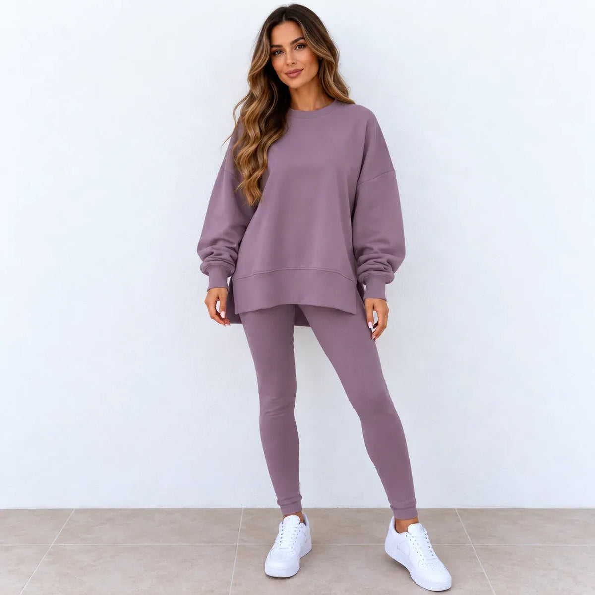 A woman modeling a matching dusty purple sweatshirt and leggings set with white sneakers, standing against a plain white wall.