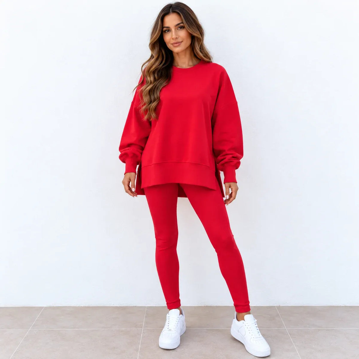 A woman modeling a matching red sweatshirt and leggings set with white sneakers, standing against a plain white wall.