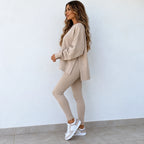 A woman modeling a matching beige lounge set consisting of an oversized long-sleeve sweatshirt and high-waisted leggings, paired with white sneakers, standing against a plain white wall.
