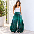 A woman modeling a pair of wide-leg harem pants in a green and teal abstract print, paired with a white tank top, standing against a white wall.