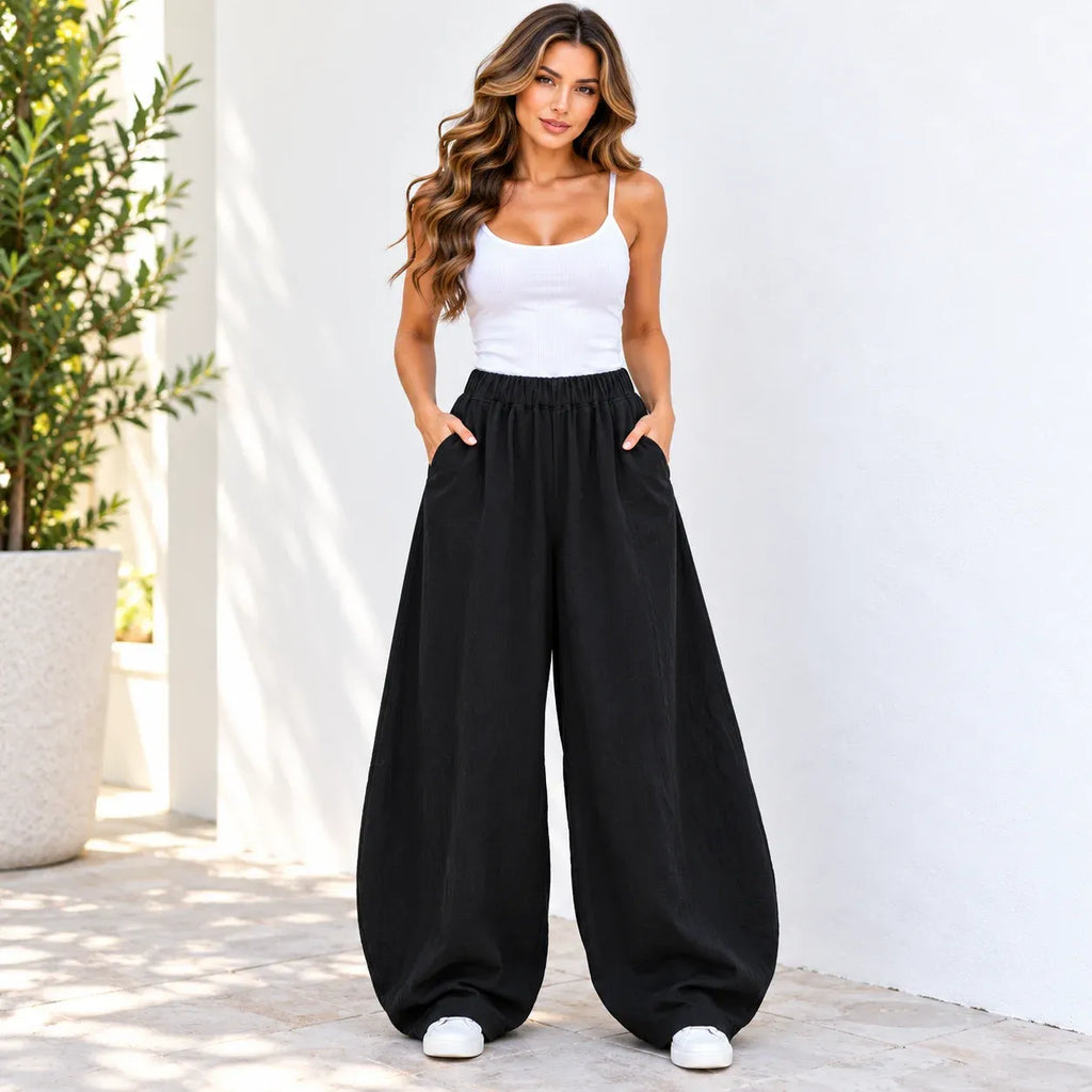 A woman stands outdoors against a white wall wearing loose black wide-leg pants with a white spaghetti-strap tank top and white sneakers.