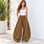 A woman stands outdoors wearing loose, balloon-style brown pants with a white camisole and beige flats, posed against a white wall with a potted plant on the left.