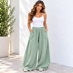 A woman modeling a pair of wide-leg sage green pants with a white camisole top, standing against a white wall with a potted plant on the left.