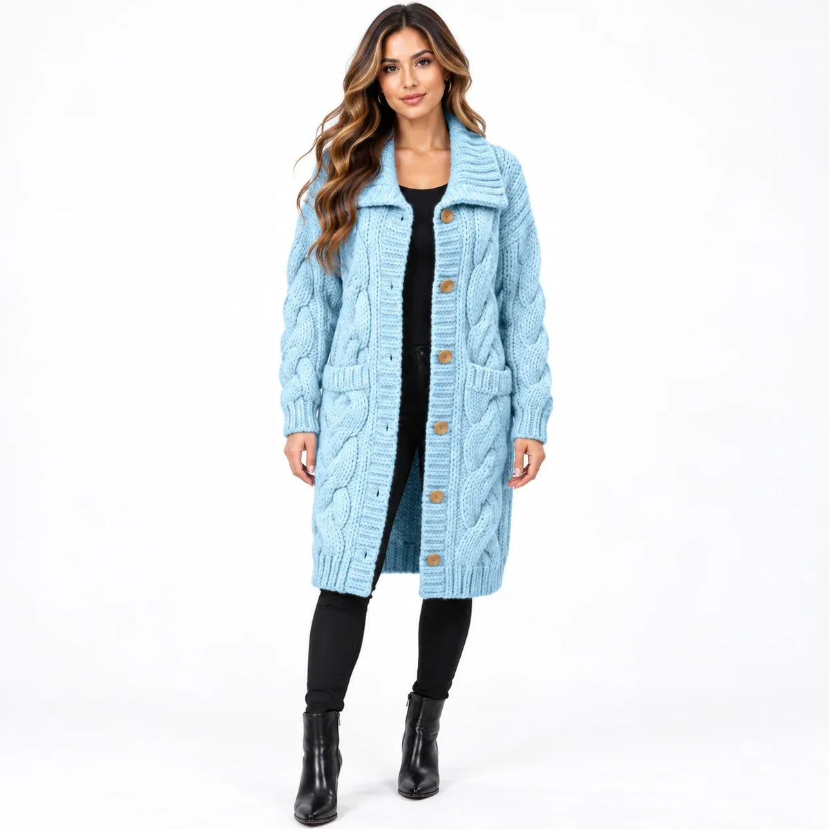 A woman modeling a light blue cable-knit cardigan with brown buttons, worn over a black dress and black boots, against a plain white studio background.