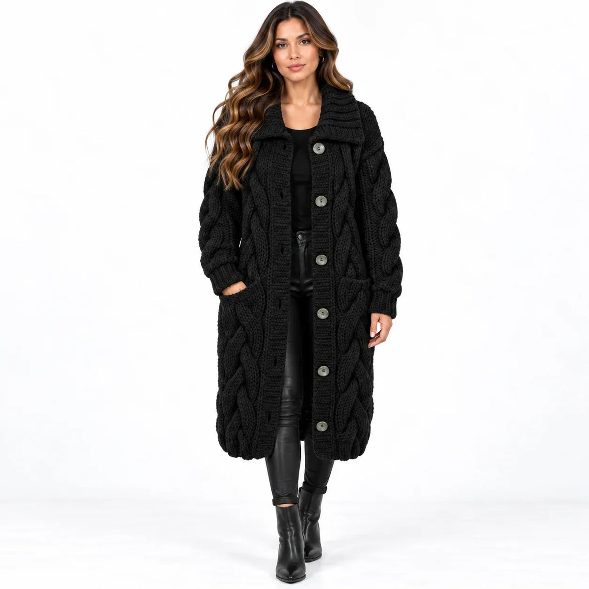 A woman modeling a long black cable-knit coat with large silver buttons, worn over a black outfit with black leather-look pants and black ankle boots, against a plain white studio background.