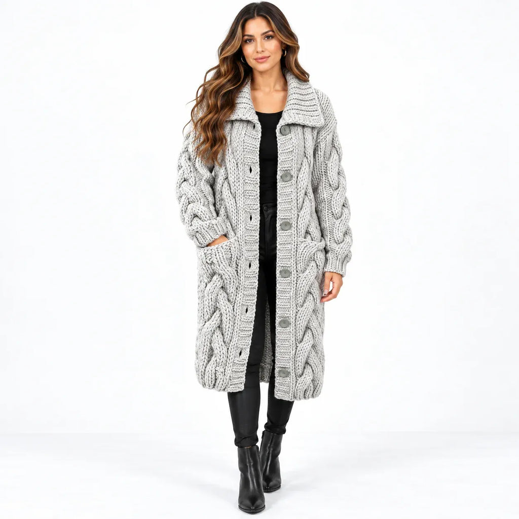 A woman modeling a long, chunky cable-knit cardigan in light gray. The cardigan features a large collar, front button closure, and side pockets, styled over a black top, black pants, and black ankle boots against a plain white studio background.
