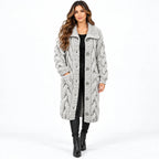 A woman modeling a long, chunky cable-knit cardigan in light gray. The cardigan features a large collar, front button closure, and side pockets, styled over a black top, black pants, and black ankle boots against a plain white studio background.