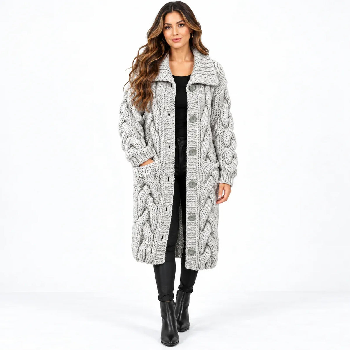 A woman modeling a long, chunky cable-knit cardigan in light gray. The cardigan features a large collar, front button closure, and side pockets, styled over a black top, black pants, and black ankle boots against a plain white studio background.
