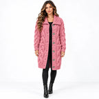 A woman modeling a pink cable-knit button-front coat with large front pockets, worn over a black outfit and black ankle boots against a plain white studio background.