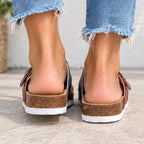 A close-up rear view of a person wearing slip-on wedge sandals with cork-look midsoles, white soles, and dark straps with red and blue side stripes, paired with frayed blue jeans.