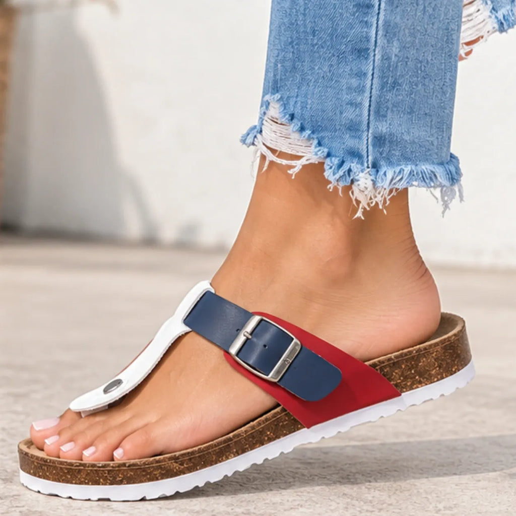 A close-up of a person's foot wearing a red, navy blue, and white thong sandal with a cork-look midsole and a silver buckle, paired with frayed blue jeans.