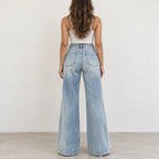 A woman viewed from behind wearing light-wash wide-leg denim jeans and a white ribbed tank top, standing against a plain off-white wall.