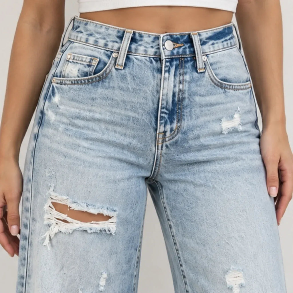 Light wash blue distressed denim jeans with ripped details on the thighs, shown on a model from the waist down against a plain light background.