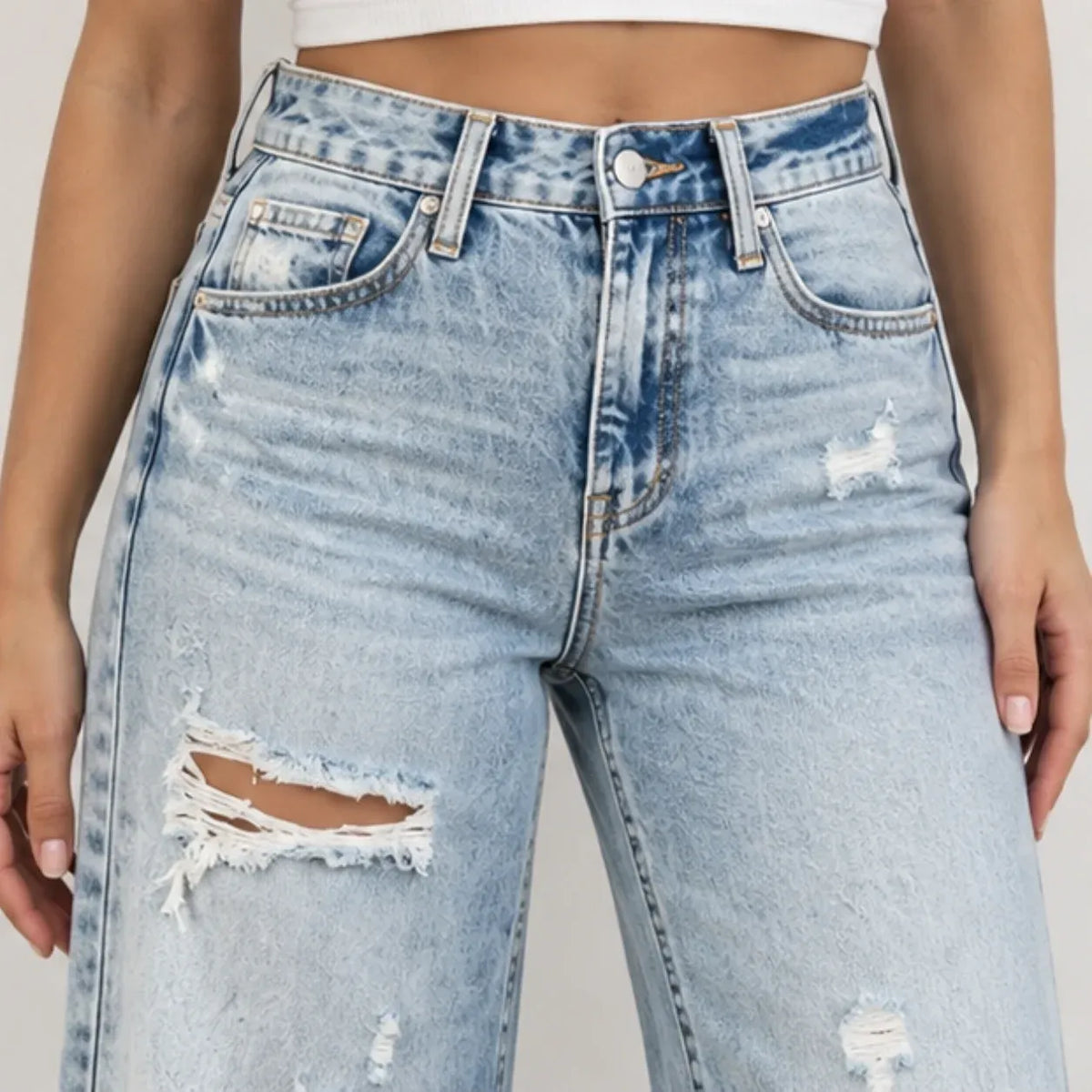 Light wash blue distressed denim jeans with ripped details on the thighs, shown on a model from the waist down against a plain light background.