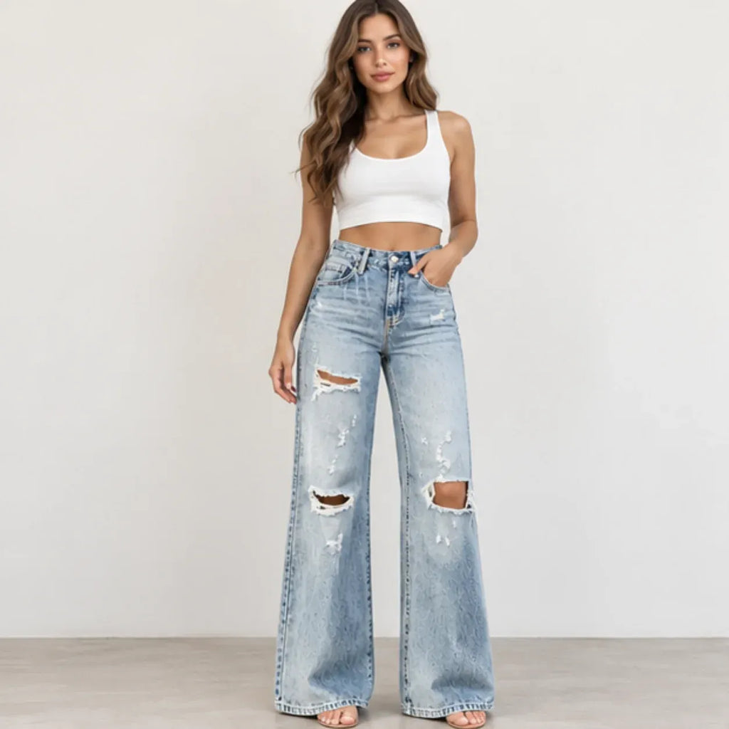 A woman modeling light-wash, wide-leg distressed jeans with ripped details on the thighs and knees, paired with a white crop top, standing against a plain off-white wall.