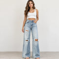 A woman modeling light-wash, wide-leg distressed jeans with ripped details on the thighs and knees, paired with a white crop top, standing against a plain off-white wall.