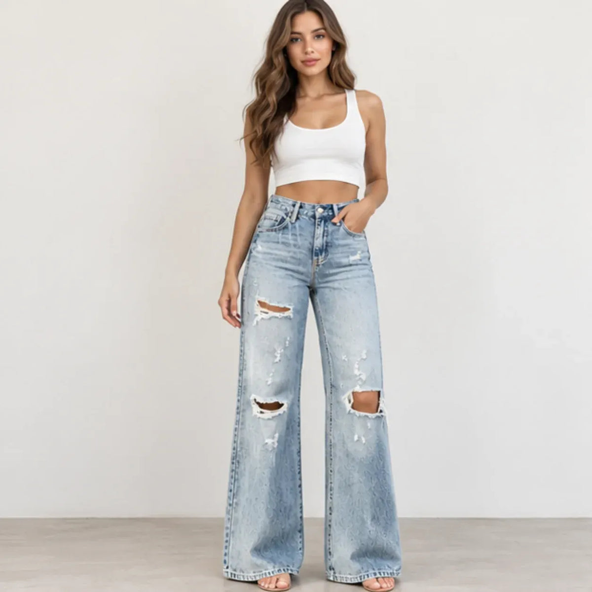 A woman modeling light-wash, wide-leg distressed jeans with ripped details on the thighs and knees, paired with a white crop top, standing against a plain off-white wall.