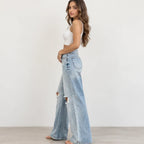 A woman modeling light-wash wide-leg distressed jeans with a white crop top, standing against a plain white wall.