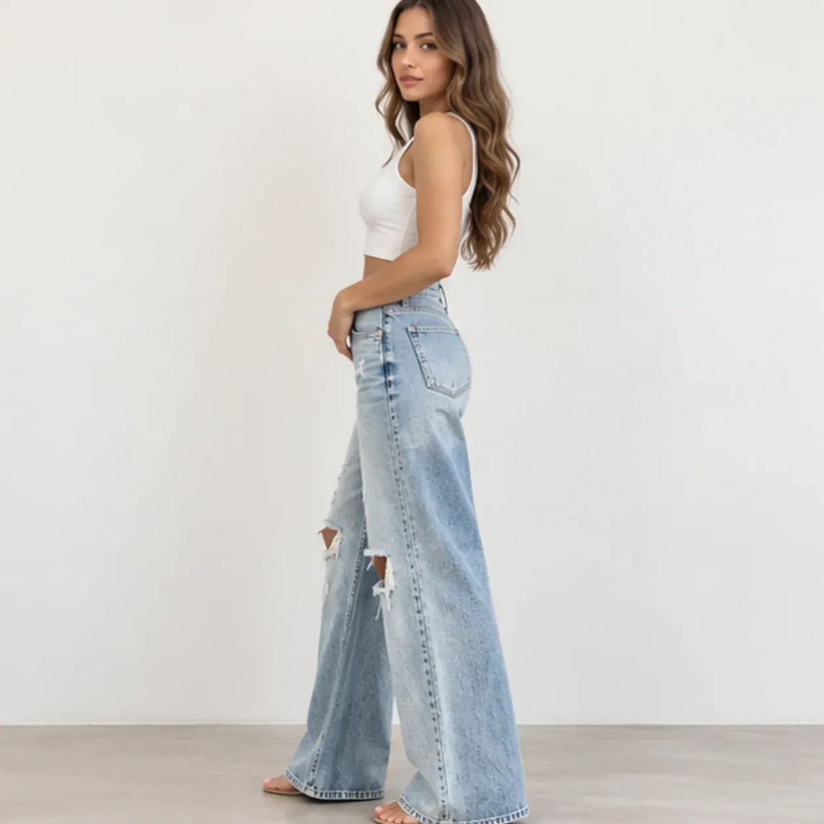 A woman modeling light-wash wide-leg distressed jeans with a white crop top, standing against a plain white wall.