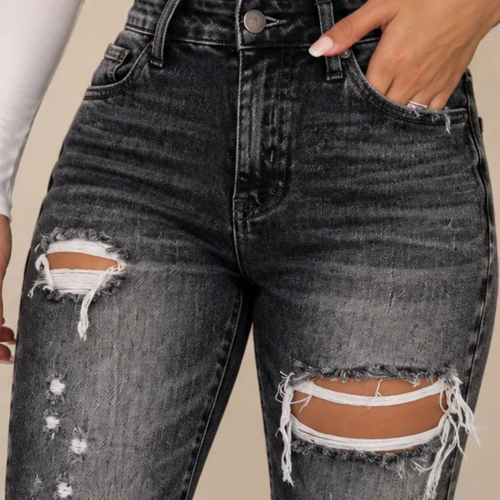 Close-up of a person wearing dark gray distressed denim jeans with ripped details and frayed edges, showing the front pocket and hand in the pocket.