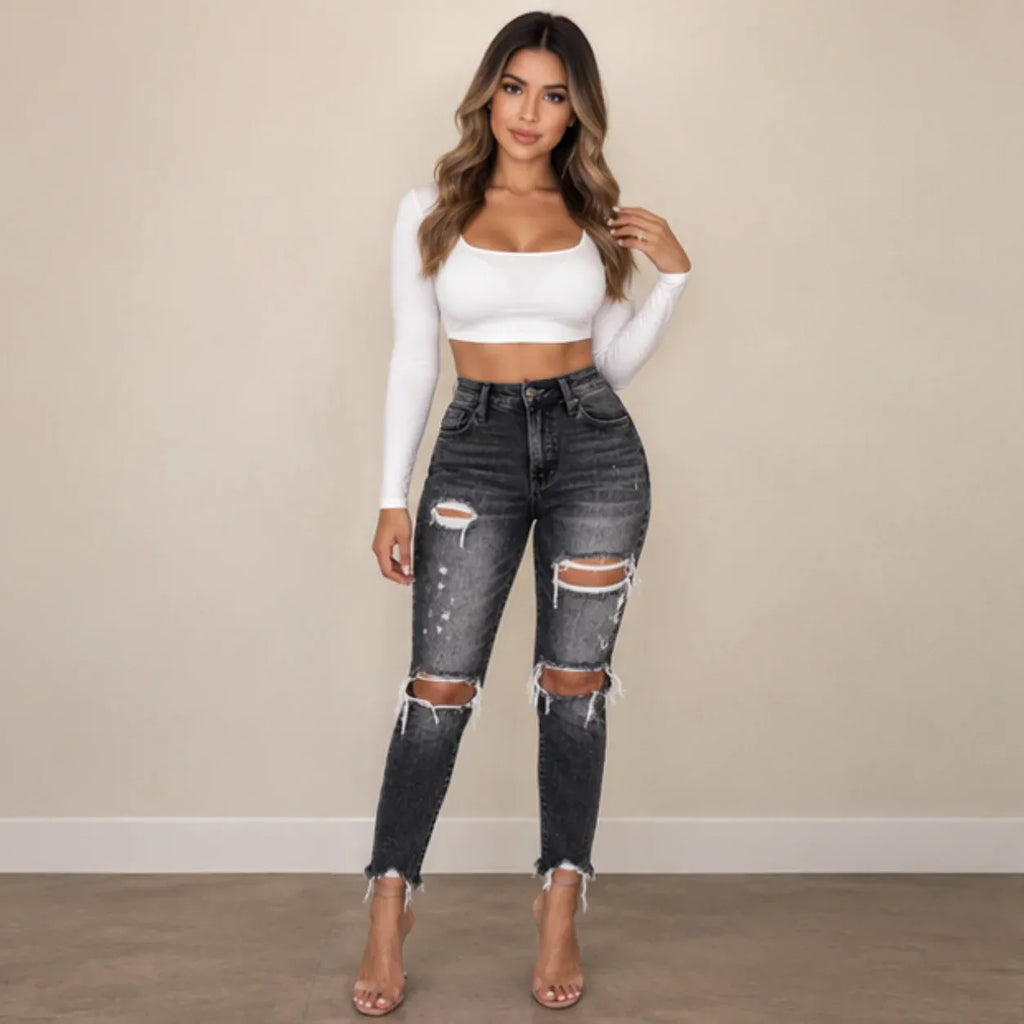 A woman modeling a pair of dark gray distressed skinny jeans with ripped knees, paired with a white long-sleeve crop top and clear high-heeled sandals, standing against a beige wall.