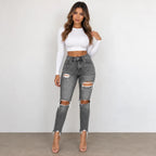 A woman modeling a white long-sleeve crop top and distressed gray skinny jeans with ripped knees, standing against a plain light gray wall.