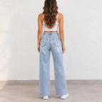 A woman viewed from behind wearing light-wash wide-leg denim jeans, a white sleeveless crop top, and white sneakers against a plain off-white wall.