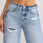 Close-up of a person wearing light-wash blue distressed denim jeans with ripped details and a faded wash, shown from the waist to mid-thigh against a plain light background.
