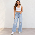 A woman modeling light-wash, wide-leg distressed jeans with a white crop top and white sneakers, standing against a plain off-white wall.
