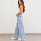 A woman wearing light-wash wide-leg distressed jeans, a white crop top, and white sneakers, standing in profile against a plain off-white wall.