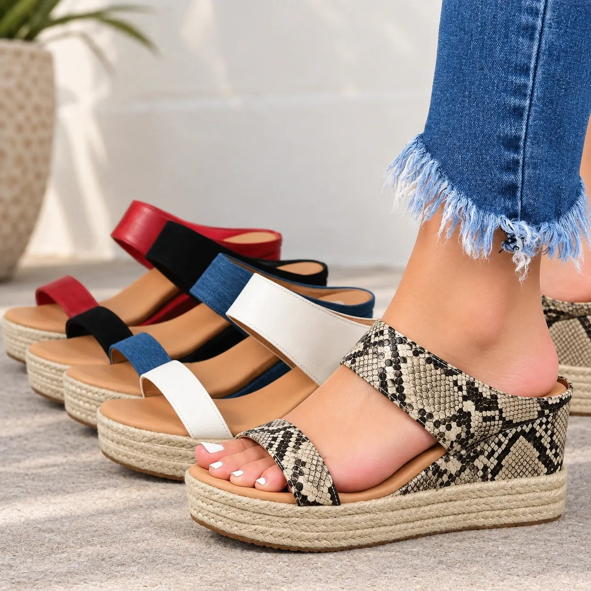 A person wearing a platform wedge sandal with a snakeskin pattern, standing outdoors next to three other color options of the same sandal lined up on a stone surface.