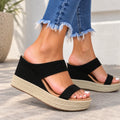 A pair of women's black wedge slide sandals with espadrille-style woven platform soles, shown on a model wearing frayed blue jeans in an outdoor setting.