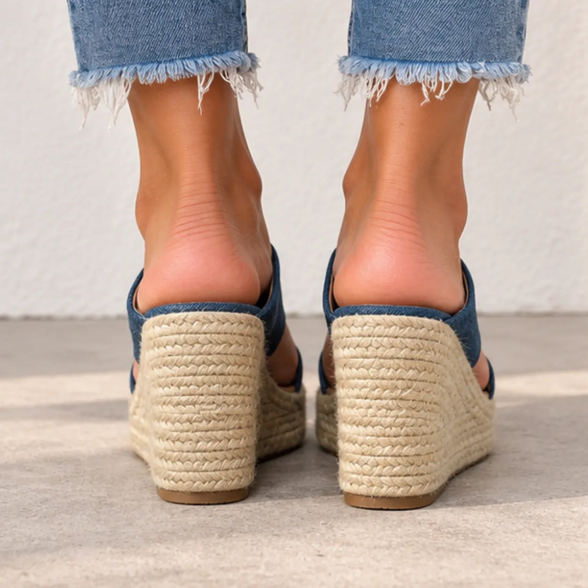 Rear view of a person wearing blue denim wedge mule sandals with thick braided jute espadrille heels and frayed hems on light blue jeans, standing on a concrete floor against a white wall.