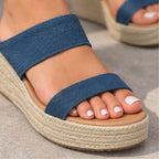 A close-up of a person's foot wearing a blue denim-look espadrille sandal with a braided jute platform sole and a tan footbed, photographed on a neutral gray surface.