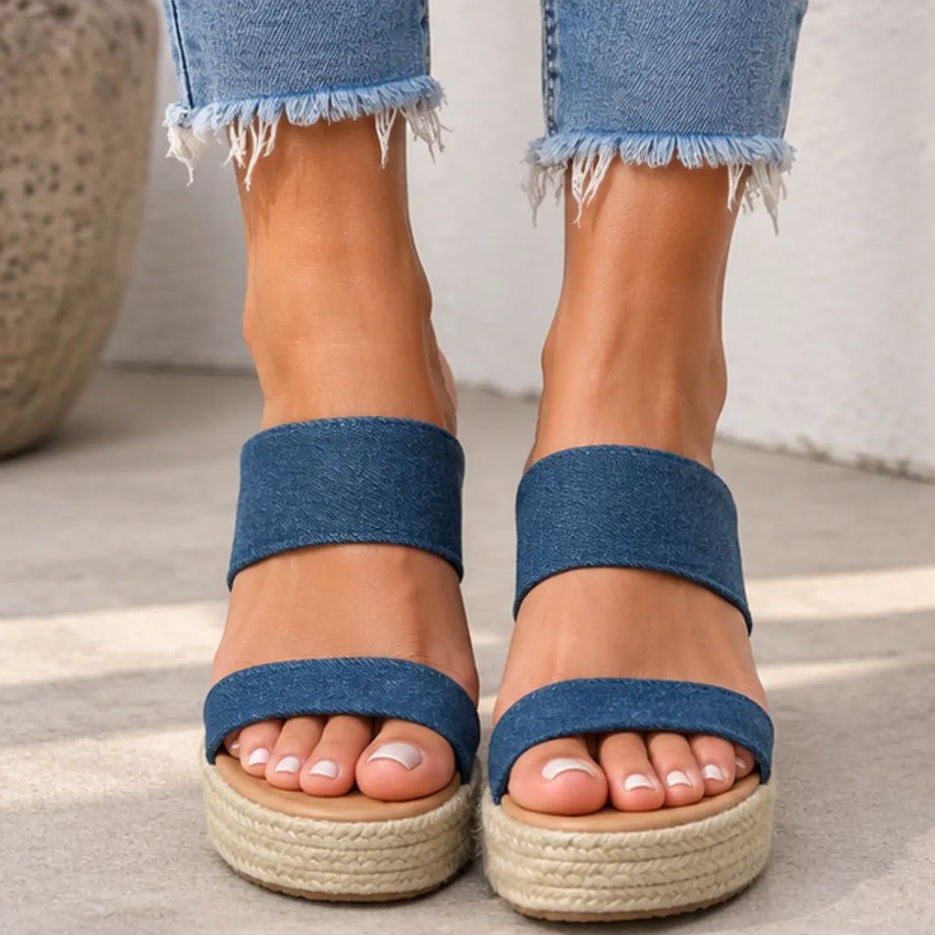 A close-up of a person wearing blue denim wedge sandals with espadrille-style woven platform soles, shown from the feet up to the jeans hems.