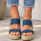 A close-up of a person wearing blue denim wedge sandals with espadrille-style woven platform soles, shown from the feet up to the jeans hems.