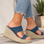 A person wearing blue denim slide wedge sandals with thick braided jute wedge heels, shown from the calves down in an outdoor setting.