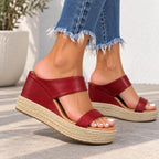 A pair of women's wedge espadrille sandals in burgundy red faux leather with braided jute platform soles, shown on a model's feet wearing frayed blue jeans.
