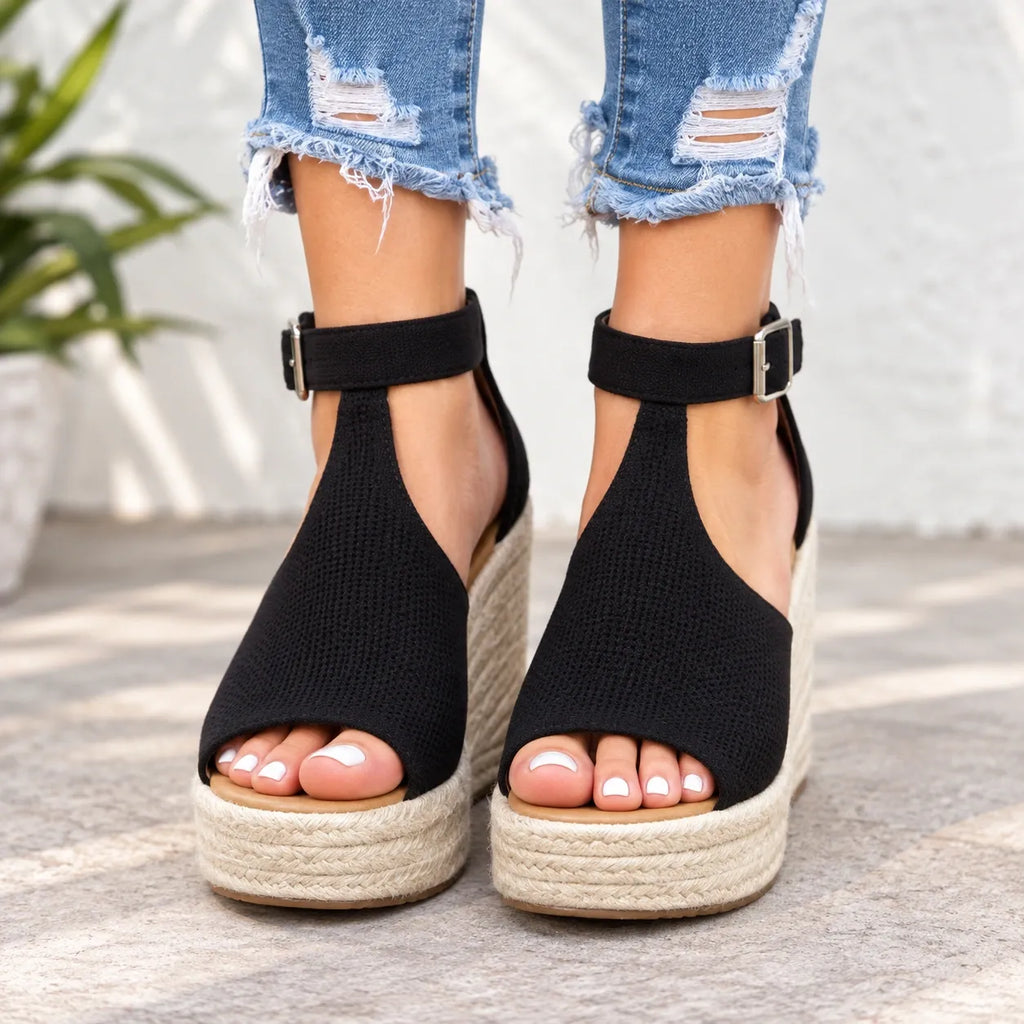 A pair of women's black woven espadrille wedge sandals with ankle straps and silver buckles, shown on a model wearing distressed blue jeans.