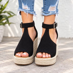 A pair of women's black woven espadrille wedge sandals with ankle straps and silver buckles, shown on a model wearing distressed blue jeans.