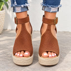A pair of women's brown espadrille wedge sandals with perforated uppers and adjustable ankle straps, shown on a model wearing frayed blue jeans.