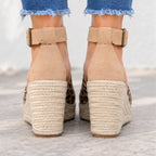 Rear view of a person wearing beige espadrille wedge sandals with leopard print sides and ankle straps, paired with frayed blue jeans on a light concrete surface.