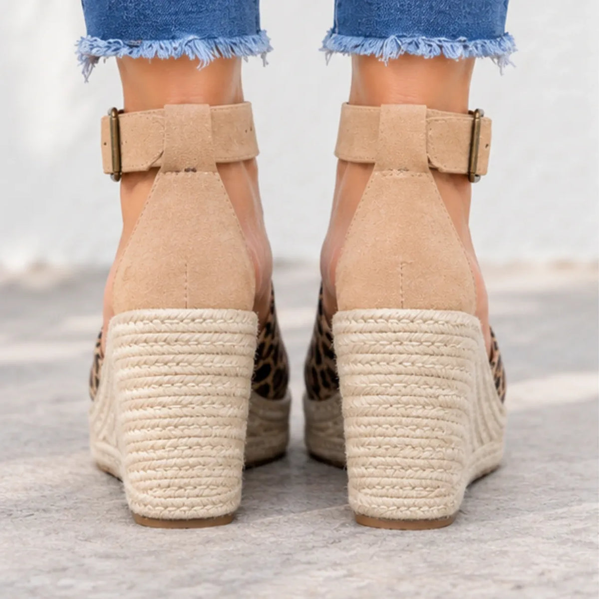 Rear view of a person wearing beige espadrille wedge sandals with leopard print sides and ankle straps, paired with frayed blue jeans on a light concrete surface.